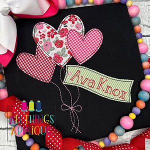 May include: Black fabric with three heart-shaped appliqués in floral and gingham patterns. A banner with the name "Ava Knox" is attached to the hearts. The design includes a bow and stitched details. The image is framed with colorful beads and bows.