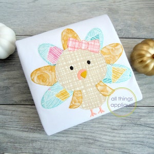 May include: A white t-shirt with a colorful patchwork turkey applique. The turkey has a pink bow and a plaid body. The text "all things applique" is visible in the lower right corner.