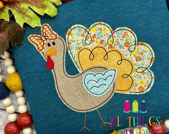 May include: A teal-coloured shirt with a turkey applique. The turkey has a bow, floral tail feathers, and a blue wing. The turkey's body is beige, with a red wattle. The text "ALL THINGS APPLIQUE" is visible in the lower right corner.