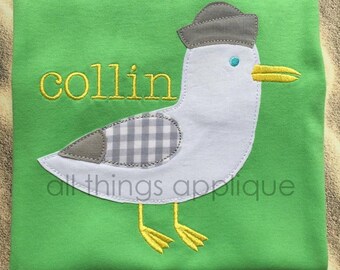 Può includere: Una maglietta verde con un'applicazione di gabbiano bianco e grigio. Il gabbiano indossa un cappello grigio e ha un becco giallo. Il nome "collin" è ricamato in giallo sulla maglietta.