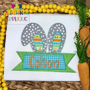 Op de afbeelding: Een wit T-shirt met een grijze en witte konijnen-applique met een blauw-groen geruit banner waarop "Colson" staat.