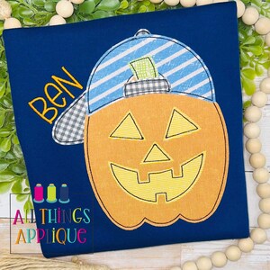 May include: A blue shirt with an orange pumpkin wearing a blue and white striped baseball cap with a plaid brim. The pumpkin has a yellow smiling face with a black outline. The name "BEN" is embroidered in yellow on the shirt.