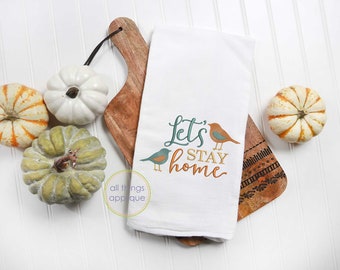 May include: White kitchen towel with the text "Let's Stay Home" in green and orange lettering with two small birds embroidered on the towel. The towel is on a wooden cutting board with a white background.