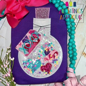 May include: Purple shirt with a white bottle filled with colorful hearts and glitter. The bottle has a pink and blue tag that says "XOXO".