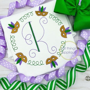 May include: A white onesie with a green and gold embroidered letter K surrounded by a wreath of purple and gold Mardi Gras masks. The onesie is decorated with purple and white polka dot ribbon and green and white striped ribbon.