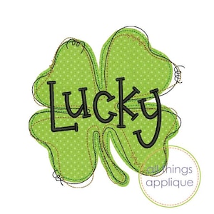May include: A green four-leaf clover design with the word "LUCKY" in black lettering. The clover has a dotted pattern and is outlined with brown and black stitching. The image has a white background.