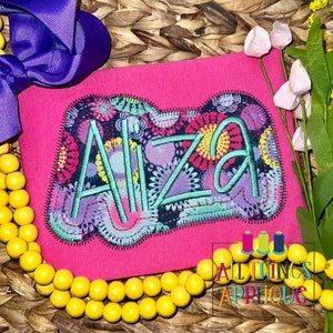 May include: A pink t-shirt with a colorful embroidered patch featuring the name "Aiza" in a whimsical font. The patch has a floral design with a black background. The t-shirt is surrounded by yellow beads and a purple bow.