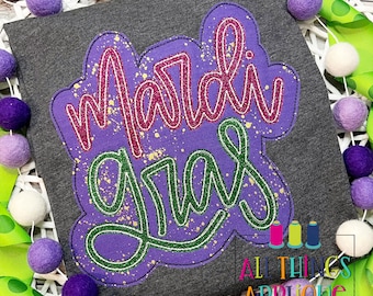 May include: Purple and green "Mardi Gras" lettering with gold glitter accents on a gray shirt.