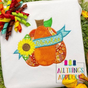May include: A personalized pumpkin applique with a sunflower and a blue ribbon with the name "Charlie" embroidered on it. The applique is made of fabric and has a patchwork design.