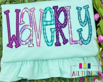Op de afbeelding: Een lichtgroen shirt met de naam "WAVERLY" geborduurd in verschillende patronen en kleuren, waaronder paarse stippen, turquoise ruitjes en bloemmotieven. Het shirt heeft een gefranjerde zoom en de tekst "ALL THINGS APRIQUE" rechtsonder.