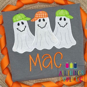 May include: A gray t-shirt with three white ghosts wearing hats. The ghosts have black eyes and mouths. The name "Mac" is embroidered in orange below the ghosts. The text "All Things Applique" is embroidered in the bottom right corner.