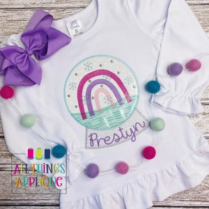 May include: A white long-sleeved shirt with a glittery snow globe applique featuring a rainbow and snowflakes. The name "Prestyln" is embroidered below the snow globe. The shirt has a ruffled hem and is decorated with a string of colorful pom-poms.
