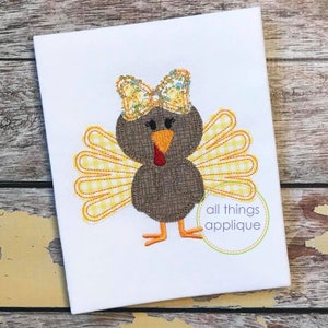 Könnte beinhalten: Gestickte Applikation eines braunen Truthahns mit gelben und weißen gemusterten Flügeln und einer Blumenschleife. Der Truthahn hat einen roten Schnabel und schwarze Augen. Das Design ist auf einem weißen Stoffhintergrund. Der Text "all things appliqué" befindet sich in einem grünen Kreis.
