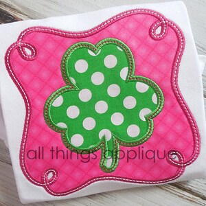 May include: A green shamrock with white polka dots is appliqued onto a pink fabric background. The shamrock is surrounded by a pink stitched frame with a scalloped edge. The text "all things applique" is visible at the bottom of the image.