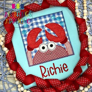 Peut inclure: Un carré de tissu vichy bleu et blanc avec une applique de crabe rouge. Le crabe a de grands yeux blancs, un sourire rouge et des pinces rouges. Le nom "Richie" est brodé en rouge sous le crabe.