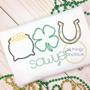 May include: A white t-shirt with green embroidered shamrocks, a gold pot of gold, and a horseshoe. The text "Sawyer" is embroidered in green below the shamrocks. The shirt is on a white wooden surface with green and gold beads.