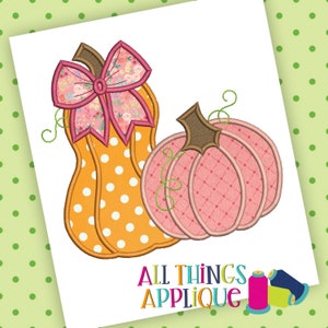 Puede incluir: Dos calabazas bordadas con un lazo rosa. La calabaza más grande es naranja con lunares blancos y la calabaza más pequeña es rosa con un patrón de diamantes. El lazo es rosa con un estampado floral.