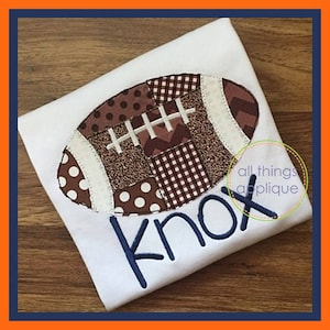 May include: A white t-shirt with a brown and white patchwork football applique and the name "Knox" embroidered in blue thread. The shirt is on a brown wooden surface. The text "all things applique" is visible in a circle on the shirt.