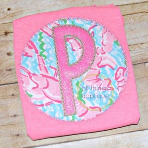May include: Pink t-shirt with a round applique featuring a pink letter P with silver glitter on a blue and pink floral pattern. The applique is surrounded by a white stitch. The text "all things applique" is visible in the lower right corner of the image.