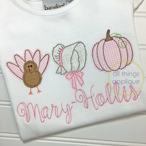 May include: White t-shirt with embroidered appliqués of a turkey, bonnet, and pumpkin. The turkey and pumpkin have pink gingham details. The name "Mary-Hollis" is embroidered in pink script. The brand name "barefoot" is visible.
