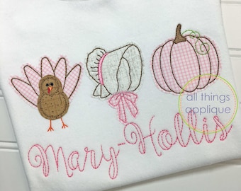 May include: White t-shirt with embroidered appliqués of a turkey, bonnet, and pumpkin. The turkey and pumpkin have pink gingham details. The name "Mary-Hollis" is embroidered in pink script. The brand name "barefoot" is visible.