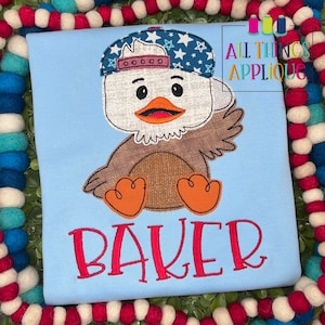 Könnte beinhalten: Eine gestickte Applikation eines Adlers, der eine rot-weiß-blaue Baseballkappe mit Sternen trägt. Der Adler sitzt mit ausgebreiteten Flügeln. Der Name "BAKER" ist in Rot unter dem Adler gestickt.