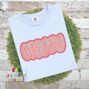 Puede incluir: Una camiseta azul claro con la palabra "Granny" bordada en rosa y azul en una fuente de burbujas.