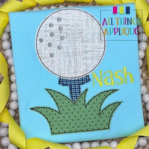 May include: A light blue t-shirt with an embroidered golf ball, green grass, and the name "Nash" in yellow thread.