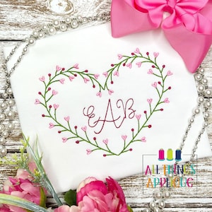 May include: A white shirt with a heart-shaped wreath of green leaves and pink flowers embroidered on it. The letters "AB" are embroidered in the center of the wreath in a cursive font.