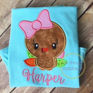May include: A light blue t-shirt with an embroidered brown turkey with a pink bow and green and orange feathers. The name "Harper" is embroidered in pink below the turkey.