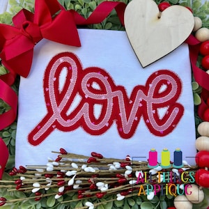 Könnte beinhalten: Rote und rosafarbene gestickte "Love" Applikation mit kleinen rosa Herzen. Die Applikation befindet sich auf einem weißen Stoffhintergrund.