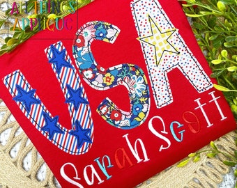 May include: Red t-shirt with the word "USA" in large, colourful letters. The letters are made of fabric scraps with stars and stripes patterns. The name "Sarah Scott" is embroidered in red, blue, and pink below the word "USA".