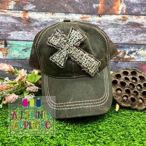 May include: A brown and black mesh trucker hat with a distressed white and brown cross applique. The hat has a white stitching detail around the brim.