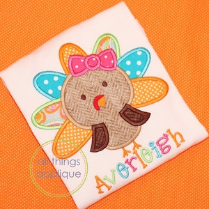 May include: A white t-shirt with an embroidered turkey design. The turkey has a brown body, a pink bow, and a colorful tail. The name "AveriLeigh" is embroidered in colorful letters below the turkey.
