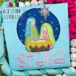 May include: A light blue t-shirt with a colorful embroidered nativity scene and the name "Stella" in pink embroidery.