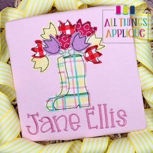 Può includere: Una maglietta rosa chiaro con un ricamo di un bouquet di tulipani in stivali di gomma a quadri. I tulipani sono rossi, viola e gialli. Il nome "Jane Ellis" è ricamato sotto. Il testo "ALL THINGS APPLIQUE" è in alto a destra.