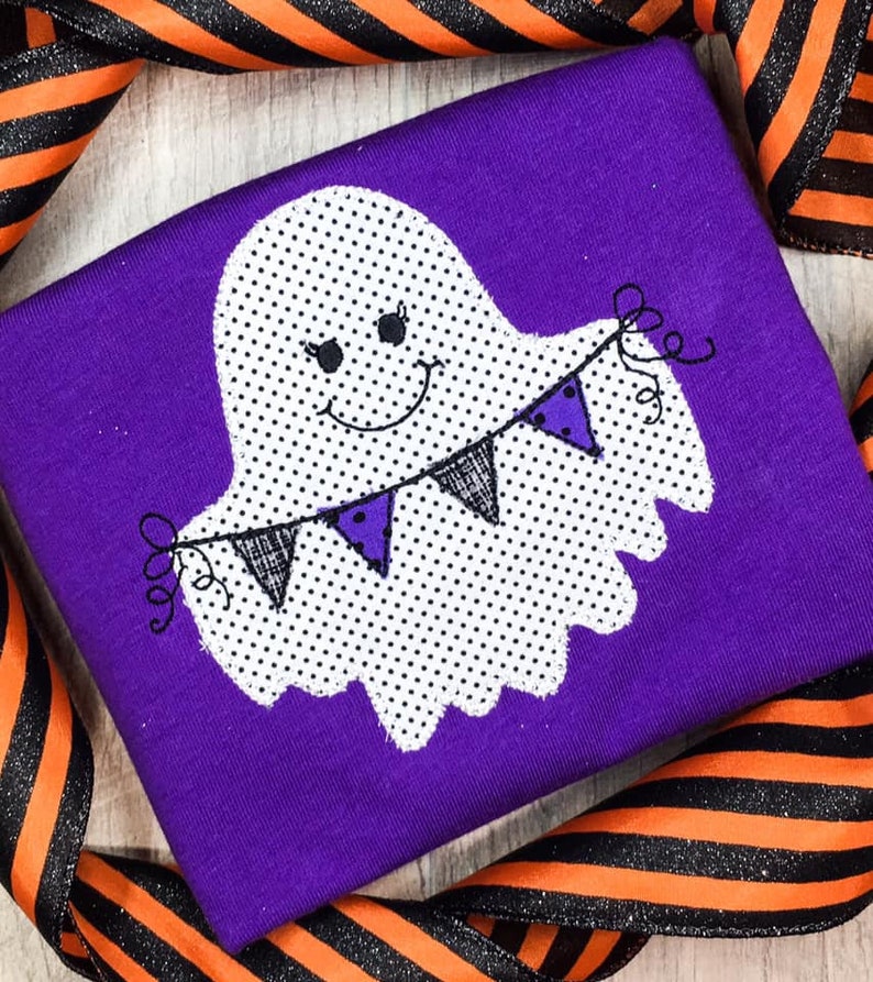May include: A purple fabric with a white ghost holding a string of black and purple pennants. The ghost has black eyes and a smiling mouth. The ghost is covered in white polka dots.