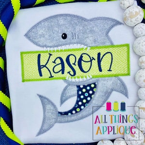 May include: A gray shark applique with a green and white chevron banner that says "Kason". The shark has a blue and white polka dot tail.