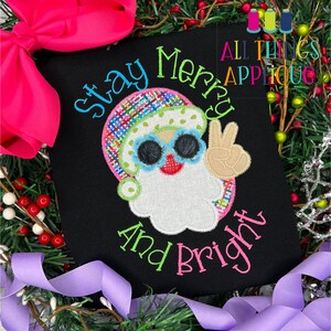 May include: A black t-shirt with a colorful applique of a Santa Claus wearing sunglasses and giving the peace sign. The text "Stay Merry and Bright" is embroidered in green and pink on the shirt.