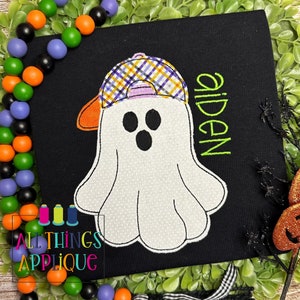 May include: A white ghost wearing a plaid baseball cap with an orange brim. The ghost has three black eyes and a black mouth. The name "aiden" is embroidered in green on the black shirt.