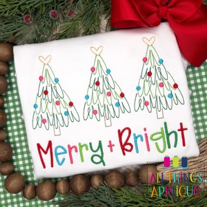 Könnte beinhalten: Weißes besticktes Weihnachtshemd mit drei grünen Weihnachtsbäumen mit roten, rosa und blauen Kugeln. Der Text "Merry + Bright" ist in bunten Buchstaben unter den Bäumen gestickt.