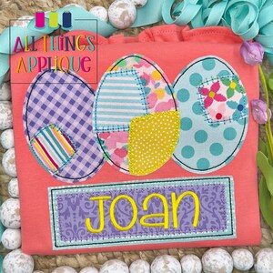 May include: A pink t-shirt with three colorful patchwork Easter eggs and a purple banner with the name "Joan" embroidered in yellow.