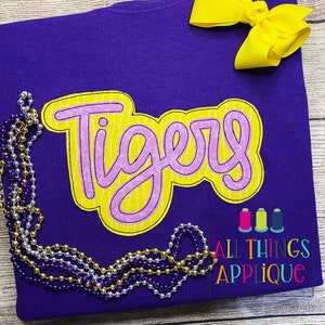 May include: Purple t-shirt with a yellow and pink applique that says "Tigers". The shirt is surrounded by gold, silver, and purple beads. The text "All Things Applique" is in the bottom right corner.