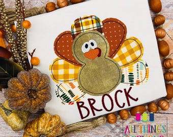 May include: A brown and gold patchwork turkey applique with googly eyes and a red beak. The turkey is on a white shirt with the name Brock embroidered in red.