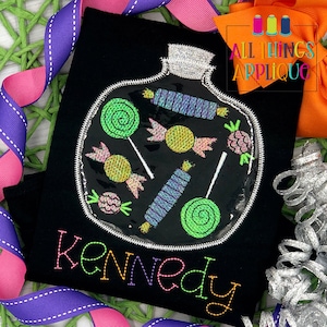 May include: A black t-shirt with a colorful embroidered design of a candy jar filled with lollipops and candy. The name "Kennedy" is embroidered in rainbow colors below the design. The shirt is decorated with purple and pink ribbon.