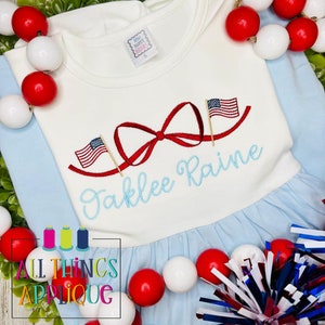 K&ouml;nnte beinhalten: Ein wei&szlig;es und blaues Babykleid mit einer roten Schleife und zwei gestickten amerikanischen Flaggen auf der Vorderseite. Der Name "Jaklee Raine" ist unter der Schleife gestickt.
