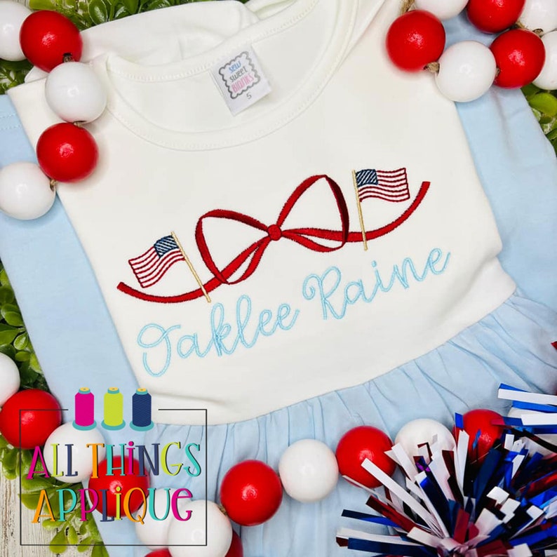 K&ouml;nnte beinhalten: Ein wei&szlig;es und blaues Babykleid mit einer roten Schleife und zwei gestickten amerikanischen Flaggen auf der Vorderseite. Der Name "Jaklee Raine" ist unter der Schleife gestickt.
