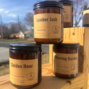 May include: Several soy candles in glass jars with black lids are stacked on a wooden shelf. The labels read "Lumber Jack" (Sandalwood), "Golden Hour" (Bergamot, Gardenia, Amber), and "Morning Garden" (Freesia, Lemon, Orange). Each candle is 8 oz.