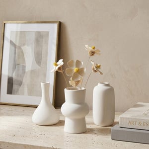 Puede incluir: Tres jarrones de cerámica blanca de diferentes formas, con flores secas, se colocan sobre una superficie de piedra. Una impresión artística abstracta enmarcada con un marco dorado y un libro titulado "ART & ESSENCE" completan la escena.