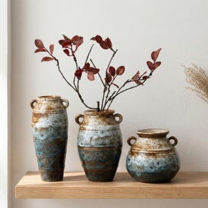 May include: Three ceramic vases with a rustic design. The vases are brown, blue, and white, with a textured finish. The tallest vase has a height of approximately 12 inches. The vases are arranged on a wooden shelf, with red branches in the center vase.